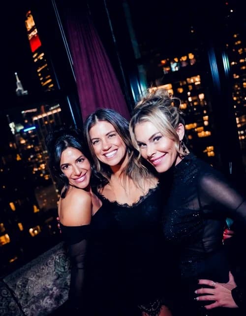 Three friends posing with drinks in hand at Suave Saturdays rooftop party, the bright city lights visible through the large windows behind them.
