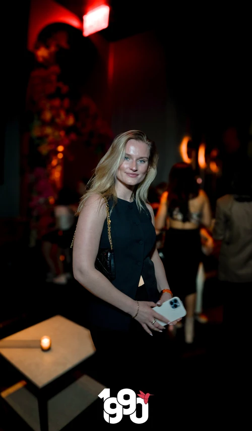 Blonde woman smiling and holding her phone, enjoying the night at Suave Saturdays with a dimly lit lounge in the background.