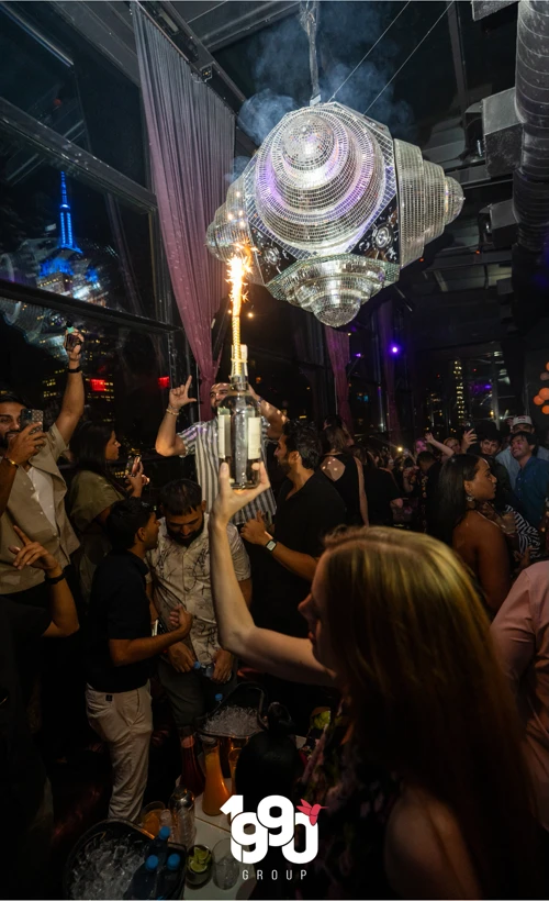 Crowded rooftop bar at Suave Saturdays in Manhattan, with a bottle of champagne being served with a sparkler and people enjoying the music and atmosphere.
