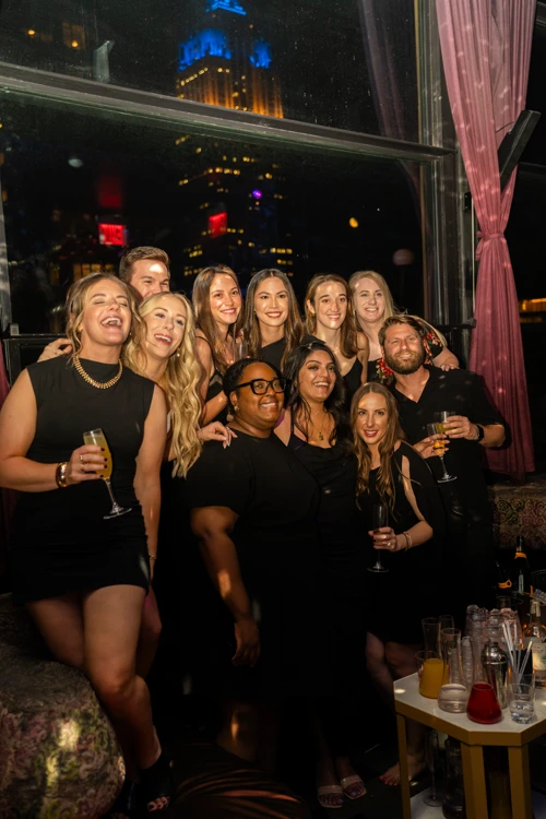 Group of friends gathered and smiling for a photo with the Manhattan skyline glowing in the background at Suave Saturdays rooftop party.