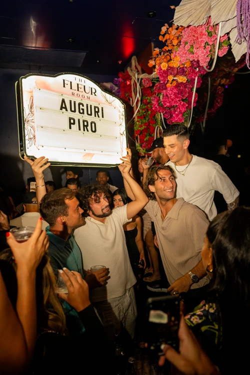 Group of friends holding up a sign that reads 'Auguri Piro' under floral decorations at Suave Saturdays rooftop party.