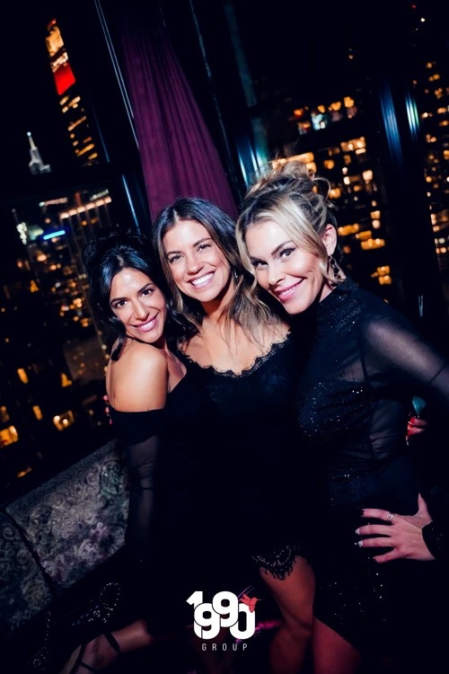 Three friends posing with drinks in hand at Suave Saturdays rooftop party, the bright city lights visible through the large windows behind them.