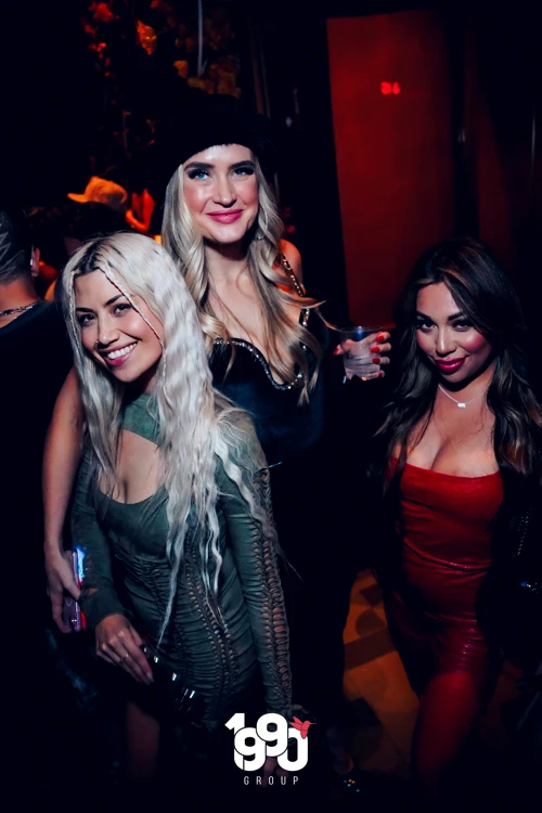 Three women dressed up for a night out, enjoying drinks and posing for a photo at Suave Saturdays, a rooftop party in Manhattan.