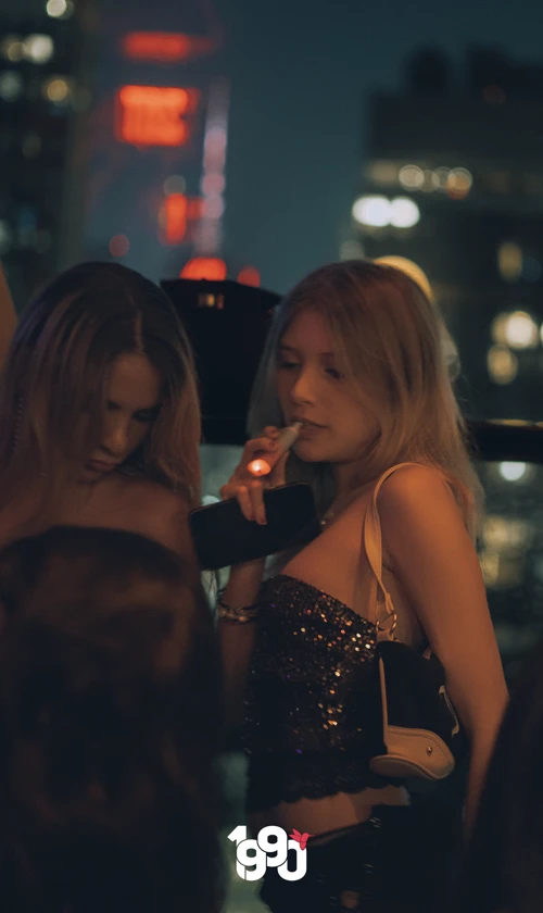 Two women chatting and vaping at Suave Saturdays rooftop event, with the city skyline visible through the windows.