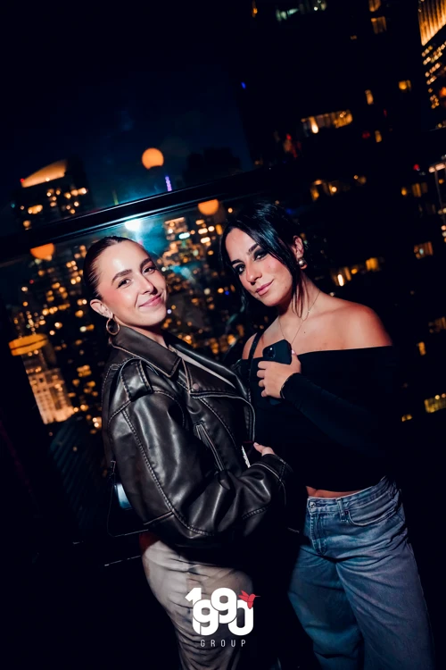 Two women smiling and posing by the window, the Manhattan skyline lit up behind them during Suave Saturdays.