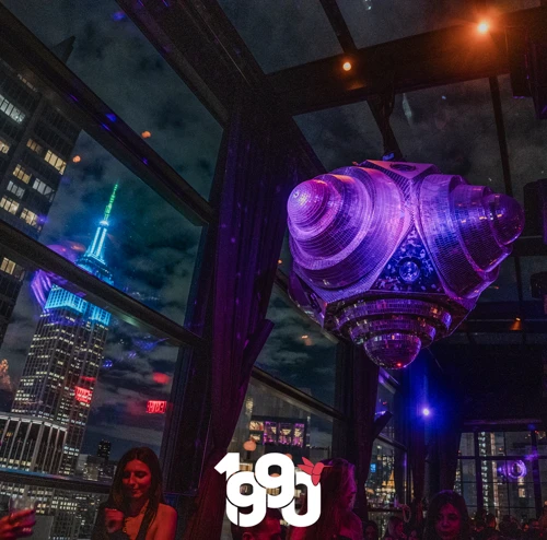 View of the Empire State Building lit up at night, with a large disco ball and partygoers in the rooftop venue of Suave Saturdays.