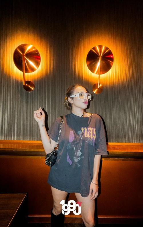 Woman posing stylishly in an oversized t-shirt and white sunglasses against a wall with modern light fixtures at Suave Saturdays.